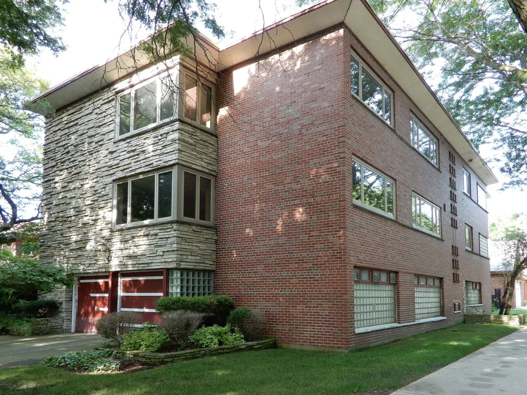 Brick and stone corner of a mid‑century modern building with large windows and a landscaped lawn nearby.